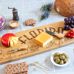 The Florida Engraved XL Charcuterie Board by Totally Bamboo, measuring 30" x 8-1/2", elegantly presents a variety of cheeses alongside a knife. It is tastefully accompanied by crackers, bread, olives, grapes, cherry tomatoes, a pepper grinder, and herbs on a subtly decorated table.