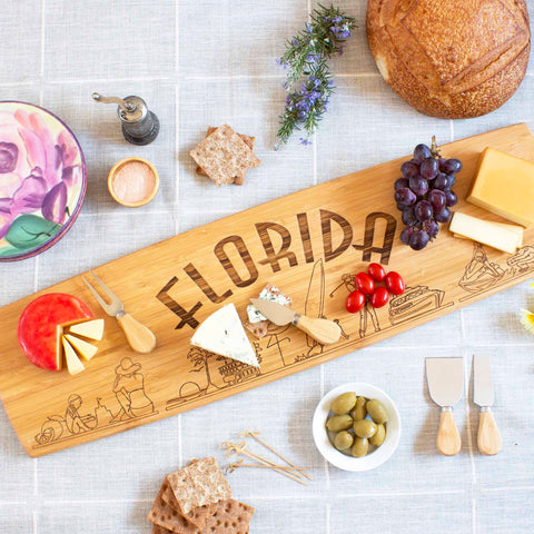 The Totally Bamboo Florida Engraved XL Charcuterie Board, measuring 30" x 8-1/2", presents a stunning wooden display with "Florida" as state artwork, elegantly arranges assorted cheeses, grapes, and tomatoes. Surrounded by crackers, olives, bread, a floral plate, an oil dispenser, and fresh rosemary sprigs on a light tablecloth—it's the quintessential cheese plate spread.