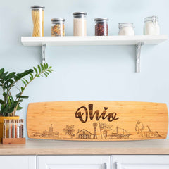 An Ohio Engraved XL Charcuterie Board by Totally Bamboo, featuring laser-engraved Ohio-themed illustrations, elegantly adorns the kitchen counter. Nearby is a potted plant, and on the white shelf above sit jars filled with pasta and spices against the light blue wall.