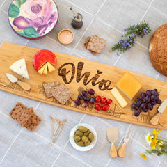 The Ohio Engraved XL Charcuterie Board by Totally Bamboo, measuring 30" x 8-1/2", showcases an assortment of cheeses, grapes, cherry tomatoes, olives, and crackers. It is paired with wooden utensils, a round floral plate, herbs, and fresh bread on a checkered tablecloth.