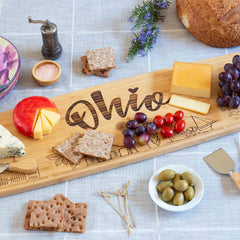 The Totally Bamboo Ohio Engraved XL Charcuterie Board, measuring 30" x 8-1/2", showcases a delightful cheese plate along with grapes, cherry tomatoes, olives, and crackers. A loaf of bread, a bowl of salt, a pepper grinder, and decorative sprigs beautifully complete the arrangement on the checkered tablecloth.