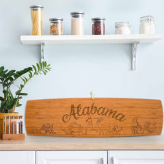 A kitchen wall showcases a laser-engraved wooden sign featuring deer and symbolic figures from Alabama. Above, shelves hold jars of pasta, grains, and legumes. A leafy plant and spice rack adorn the counter, providing a cozy backdrop for the Totally Bamboo Alabama Engraved XL Charcuterie Board measuring 30" x 8-1/2".