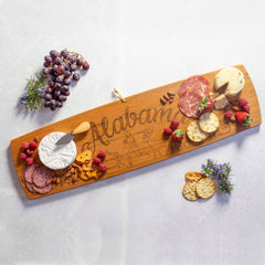 The Totally Bamboo Alabama Engraved XL Charcuterie Board, measuring 30" x 8-1/2", is elegantly arranged with a variety of cheeses, cured meats, crackers, and raspberries. Sprigs of rosemary and clusters of grapes accentuate the display on a light marble surface.