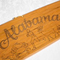 The Totally Bamboo Alabama Engraved XL Charcuterie Board, measuring 30" x 8-1/2", features the word "Alabama" elegantly engraved in cursive. Beneath this is a detailed laser engraving of a football player, a deer, and a crab, making it ideal for presenting your finest cheeses.