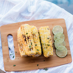 Grilled corn on the cob, adorned with herbs, is artfully presented on a Totally Bamboo cutting board from their 3-Piece Cutting Board Set. Three lime slices are placed alongside the corn, and the board is elegantly positioned atop a white cloth.