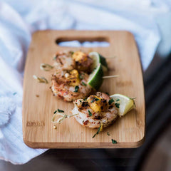 Grilled shrimp skewers, garnished with herbs and pineapple pieces, are artistically displayed on a Totally Bamboo cutting board from the 3-Piece Cutting Board Set, alongside fresh lime wedges.