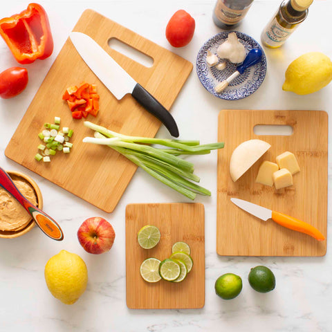 A top view of a kitchen scene features a 3-Piece Cutting Board Set by Totally Bamboo. Neatly sliced vegetables, fruits, and cheese are displayed, including red pepper, scallions, onion, lime, lemon, apple, tomatoes. Two knives with black and orange handles are also visible.