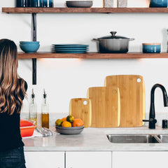 A woman with long hair stands at a kitchen counter, facing away. A Totally Bamboo 3-Piece 2-Tone Cutting Board Set made from Moso bamboo is displayed alongside her. Open shelves hold dishware and a pot above, while a bowl of fruit and bottles of oil adorn the countertop.