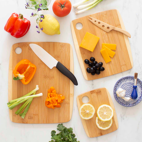 A kitchen scene featuring the Totally Bamboo 3-Piece 2-Tone Cutting Board Set crafted from Moso bamboo, displaying chopped yellow bell pepper, scallions, lemon slices, cheddar cheese, and black olives. It is surrounded by fresh vegetables, a ceramic bowl, and a knife.