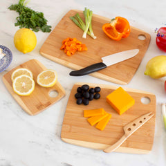 In a kitchen scene, the Totally Bamboo 3-Piece 2-Tone Cutting Board Set is featured, highlighting three Moso bamboo boards. One board holds sliced lemon, while another showcases a two-tone design with chopped bell pepper and green onions next to a knife. The third board displays black olives and cheddar slices accompanied by a cheese knife. A lemon, tomato, and parsley are arranged around them.