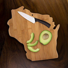 A Wisconsin Cutting Board from Totally Bamboo, measuring 13" x 12", stylishly displays a half avocado, three avocado slices, and a knife with a black handle on top of its state-shaped bamboo surface, set against a dark wooden background.