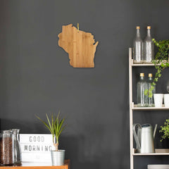 A wooden decoration in the shape of Wisconsin, specifically a 13" x 12" cutting board from Totally Bamboo, adds charm to a dark gray wall. Nearby, a shelf holds glass bottles, plants, and a watering can. On the table, a bamboo charcuterie board complements a lightbox displaying "Good Morning," with jars filled with coffee beans nearby.