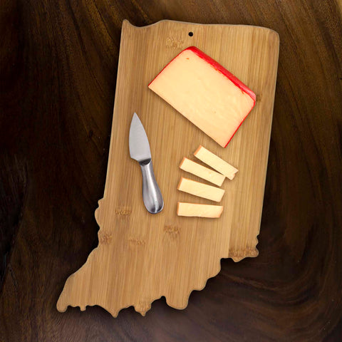 The Totally Bamboo Indiana Cutting Board, measuring 15-3/4" x 10", serves as a sturdy base for a small cheese knife, a wedge of red-rinded cheese, and four slices of cheese. This delightful charcuterie board sits on a dark wooden surface, ready to enhance your culinary display.