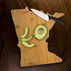 The Totally Bamboo Minnesota Cutting Board, measuring 13-1/4 x 11-3/4 inches, is adorned with sliced avocado pieces. A knife featuring a black handle rests beside the slices on a dark wooden surface, making it ideal for your charcuterie board needs.