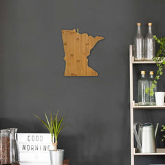 A Totally Bamboo "Minnesota" Cutting Board, measuring 13-1/4 by 11-3/4 inches, hangs on a dark gray wall. To the right, a shelf displays glass bottles and a watering can. Below, a lightbox that reads "Good Morning" sits beside a small plant in a container.