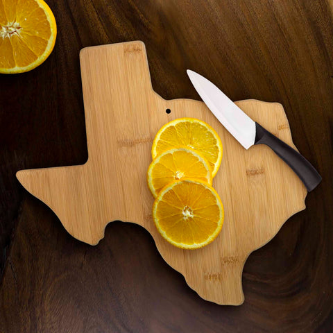 The Totally Bamboo Texas Cutting Board, measuring 14" x 13-3/8", is displayed on a dark surface. Crafted from wood, it's ideal as a charcuterie board and elegantly holds several sliced oranges next to a knife with a black handle.