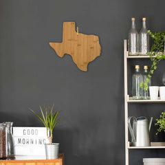 A wall displays the "Texas Cutting Board" by Totally Bamboo, measuring 14" x 13-3/8", next to a shelving unit adorned with glass bottles and plants. Beneath it, a wooden counter holds a lightbox that says "Good Morning" alongside a green plant in a pot.