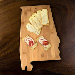 The Totally Bamboo Alabama Cutting Board, measuring 15-3/4" x 10", artfully showcases slices of bread and two open sandwiches adorned with meat and cherry tomatoes, serving as a delightful charcuterie board. This beautifully crafted wooden board is elegantly positioned on a dark wooden surface.