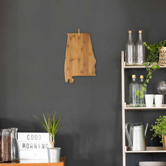 An Alabama Cutting Board, measuring 15-3/4" by 10" and crafted by Totally Bamboo, is displayed on a dark wall. Adjacent to it is a shelf adorned with glass bottles, plants, and kitchen essentials. A "Good Morning" sign rests on the counter next to a potted plant.