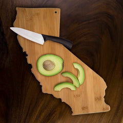 On a dark wooden table, a Totally Bamboo California Cutting Board, 14-1/4" x 11", holds a ceramic knife and a sliced avocado.