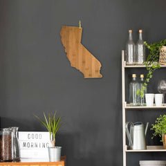 A wooden California-shaped wall decor hangs on a dark gray wall. Nearby, a shelf holds glass bottles and potted plants. A "Good Morning" sign and a watering can sit on a wooden cabinet, alongside Totally Bamboo's state-shaped bamboo cutting board, measuring 14-1/4" x 11", perfect for your next charcuterie board assembly.