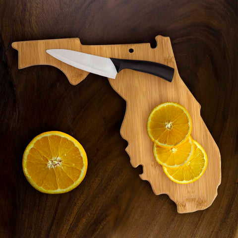 A Totally Bamboo state-shaped cutting board, designed in the shape of Florida and measuring 13-1/2" x 12-1/4", showcases sliced oranges. A knife with a black handle is placed next to the fruit, all set against a dark wood surface, making it perfect for enhancing your charcuterie board presentation.