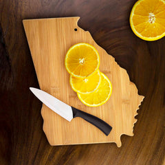 A Totally Bamboo Georgia Cutting Board, measuring 13-1/4" x 11-3/4", rests on a dark surface. The charcuterie board holds a knife with a black handle and three slices of an orange, while two additional orange halves are positioned nearby.