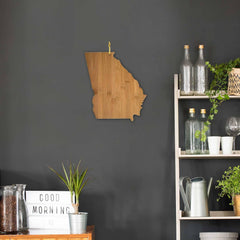 A dark wall features a Totally Bamboo Georgia Cutting Board, measuring 13-1/4" x 11-3/4", shaped like the state of Georgia. To the right, a gray shelf holds glass bottles, wine glasses, and a potted plant. Below, a small table displays a "Good Morning" sign next to another lush potted plant.