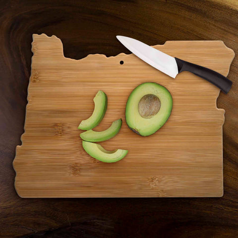 The Totally Bamboo Oregon Cutting Board, measuring 14-1/2" x 10-3/4", is ideal for serving charcuterie. It elegantly displays a halved avocado with slices, complemented by a sleek black-handled knife nearby. When not in use, this charming piece doubles as wall art, infusing rustic elegance into any kitchen space.