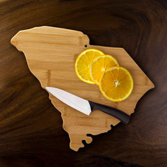 The South Carolina Cutting Board by Totally Bamboo, measuring 14-1/4" x 11", beautifully showcases three slices of orange next to a ceramic knife with a black handle, all placed against a luxurious wooden background – ideal for use as a charcuterie board or for slicing and dicing.