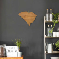 In a minimalist room with a dark grey wall, a wooden South Carolina state cutout is prominently displayed. Nearby, a shelf showcases clear glass bottles, plants, and white cups. On the table sits a small plant and the Totally Bamboo South Carolina Cutting Board (measuring 14-1/4" x 11"), alongside a lightbox displaying "GOOD MORNING.