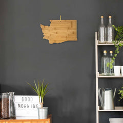 A stylish kitchen with a dark gray wall showcases the Washington Cutting Board, 15-1/2" x 10" by Totally Bamboo, shaped like Washington State. On a wooden shelf are glass bottles, plants, and a watering can. A "Good Morning" sign and potted plants decorate the wooden countertop.