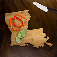 A Louisiana-shaped cutting board from Totally Bamboo, measuring 13" x 12", features vibrant sliced red bell peppers and lime wheels. Nearby, a knife with a black handle rests on the dark wooden surface, enhancing this rustic charcuterie board display.