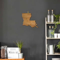 A wall decor resembling the Louisiana Cutting Board by Totally Bamboo, measuring 13" x 12", is displayed on a dark wall. Nearby, a shelf is filled with charcuterie boards, bottles, plants, and watering cans. A table showcases a letter board with "GOOD MORNING" alongside a potted plant.