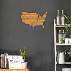 A Totally Bamboo USA Cutting Board, measuring 17-1/2" x 11", adorns the dark wall. Beside it, a shelf with glass bottles, a watering can, and potted plants enhances the room's charm. A lightbox displaying "GOOD MORNING" and a small plant rest on a wooden surface enhanced by a charcuterie board.