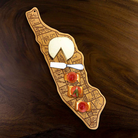 A wooden charcuterie board shaped like Manhattan showcases slices of cheese, crackers, and cherry tomatoes. The Totally Bamboo City Life Manhattan Cutting Board, measuring 7-1/4" x 22", is laser-engraved with neighborhood names such as Harlem and Hell's Kitchen in a playful map style.