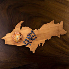 A robust Michigan Upper Peninsula Cutting Board by Totally Bamboo, measuring 17" x 9-1/2", beautifully displays a blueberry muffin and scattered blueberries on a dark wooden surface.