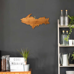 In a minimalist room with a dark gray wall, a wooden art piece shaped like an island is displayed. Below, a shelf showcases glass bottles, lush green plants, and a watering can. A durable Michigan Upper Peninsula Cutting Board from Totally Bamboo rests on the cabinet next to a box labeled "Good Morning" and a potted plant.