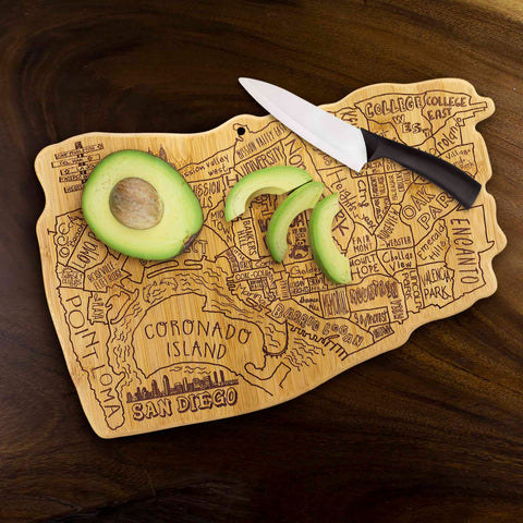 An avocado sliced open and a black-handled knife rest on the Totally Bamboo City Life San Diego Cutting Board, measuring 14-1/2" x 9-3/8", featuring city names laser-engraved on its carved wooden surface. The bamboo cutting board sits elegantly on a dark wooden table.