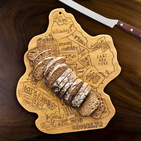 A loaf of sliced bread sits on a City Life Brooklyn Cutting Board by Totally Bamboo, measuring 13-1/2" x 11-3/4" and adorned with laser-engraved neighborhood names. A serrated knife rests on the table nearby.