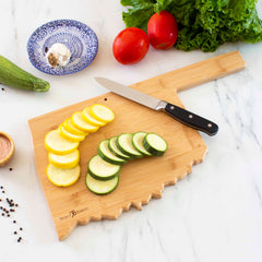 The Totally Bamboo Destination Oklahoma Cutting Board, measuring 16-3/4" x 9", holds neatly sliced yellow and green zucchini with a knife resting beside them. In the background, lettuce, tomatoes, a small bowl with garlic, and peppercorns are visible on a marble surface.