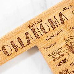 The Destination Oklahoma Cutting Board by Totally Bamboo measures 16-3/4" x 9" and is crafted from bamboo, laser-engraved with "Oklahoma," featuring towns such as Buffalo, Woodward, and Cheyenne. A small cowboy hat and lasso illustration adds charm to this unique piece.