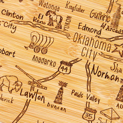 The Totally Bamboo Destination Oklahoma Cutting Board, measuring 16-3/4" x 9", artistically represents parts of Oklahoma with a stylized design. It highlights cities such as Oklahoma City, Norman, and Lawton. The bamboo surface is intricately laser-engraved with artwork featuring animals, a covered wagon, and various highways on a wood grain texture.