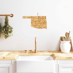 A modern kitchen features a white sink, wooden countertop, and copper faucet. Above the sink, a Totally Bamboo Destination Oklahoma Cutting Board measuring 16-3/4" x 9" hangs as a decorative piece. Dried herbs and kitchen utensils are placed on the bamboo cutting surface.