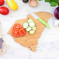 A 14" x 11-1/2" Destination South Carolina Cutting Board by Totally Bamboo, shaped like the state and featuring a bamboo surface, sits on a marble counter topped with sliced tomatoes and cucumbers. A knife with a green handle rests beside the laser-engraved artwork. Nearby are tomatoes, squash, purple onion, garlic, and herbs.