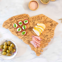 A Destination South Carolina Cutting Board by Totally Bamboo, measuring 14" x 11-1/2", is adorned with small sandwiches, sliced cheese, and ham. The cutting board features laser-engraved artwork of city names that adds a touch of charm. Nearby, a bowl of olives and a bowl of salt are placed on the marble surface.