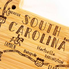 Here's a rewritten version of the sentence using the provided product data:

The Totally Bamboo Destination South Carolina Cutting Board, measuring 14" x 11-1/2", is a detailed bamboo cutting surface laser-engraved with "South Carolina" and the names of towns such as Camden, Hartsville, and Darlington. It also features an illustration of a horse and rider along with highlights of notable roads like Route 20.