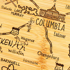 The Totally Bamboo Destination South Carolina Cutting Board, measuring 14" x 11-1/2", features laser-engraved artwork with a hand-drawn map of the state. It highlights cities like Columbia, Aiken, and Orangeburg, includes roads such as Highway 20 and landmarks like Lake Murray, and even depicts a charming horse near Wagener.