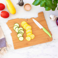 The Destination Oregon Cutting Board by Totally Bamboo, measuring 14-1/2" x 10-3/4", is elegantly displayed on a marble countertop, adorned with slices of zucchini and yellow squash. Beside it lies a knife with a green handle, accompanied by fresh tomatoes, a pepper mill, salt, and basil. A cloth towel is casually set to the side.