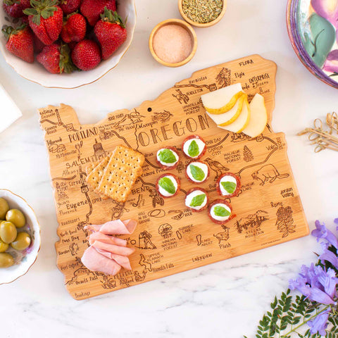 A Totally Bamboo Destination Oregon Cutting Board, measuring 14-1/2" x 10-3/4", artistically displays sliced pears, crackers, prosciutto, and snacks topped with cream cheese and herbs. Nearby are bowls of strawberries and olives on a marble countertop, accented by a purple flower.